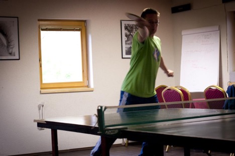 Osoba hrajúca stolný tenis v miestnosti s dreveným stolom a červenými stoličkami.