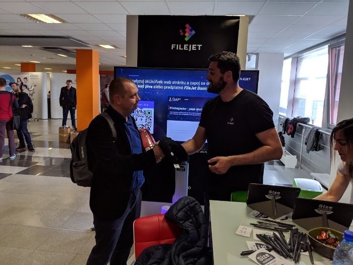 Dvaja muži si podávajú ruky na podujatí FileJet, kde prebieha prezentácia alebo diskusia o rýchlosti webu.