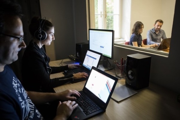 Ľudia pracujúci na počítačoch v kancelárii, dvaja v popredí so slúchadlami, dvaja v pozadí za sklom