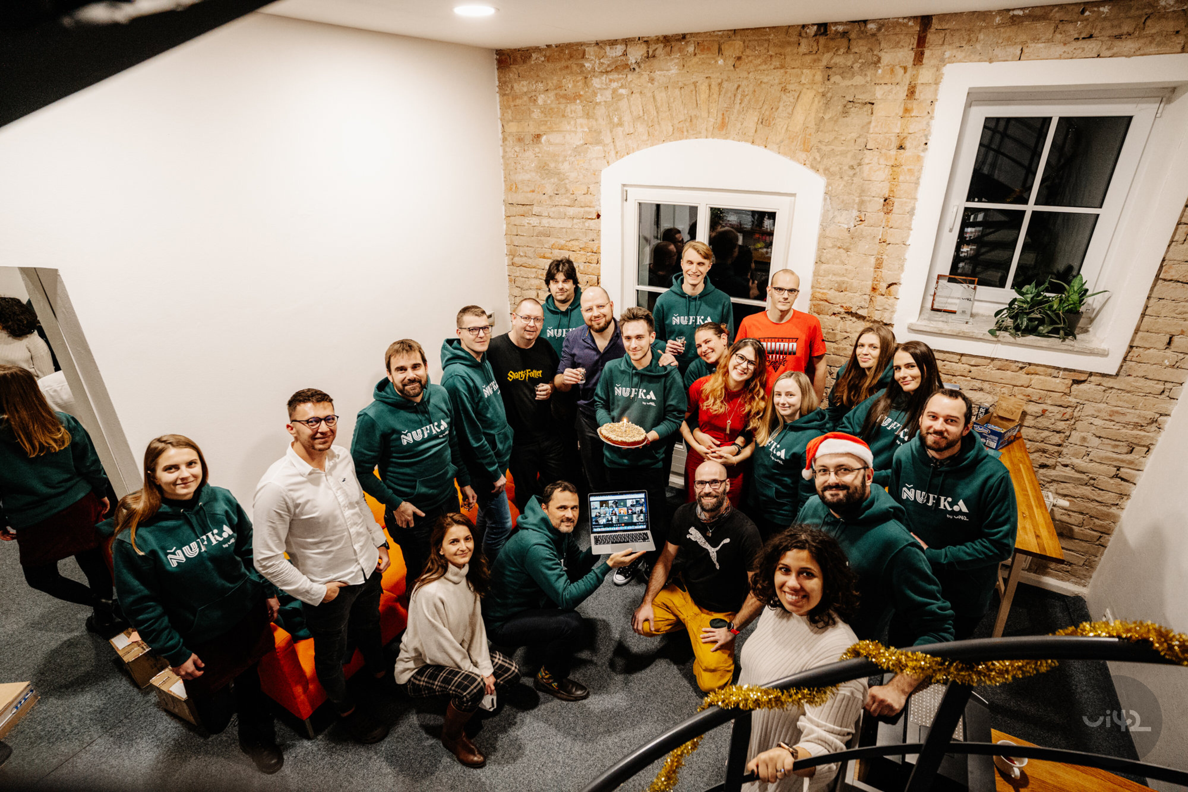 Skupina ľudí stojí a sedí v miestnosti s bielymi stenami a tehlovým múrom, niektorí majú oblečené zelené mikiny s nápisom 'Nufka', usmievajú sa a pozerajú do fotoaparátu.