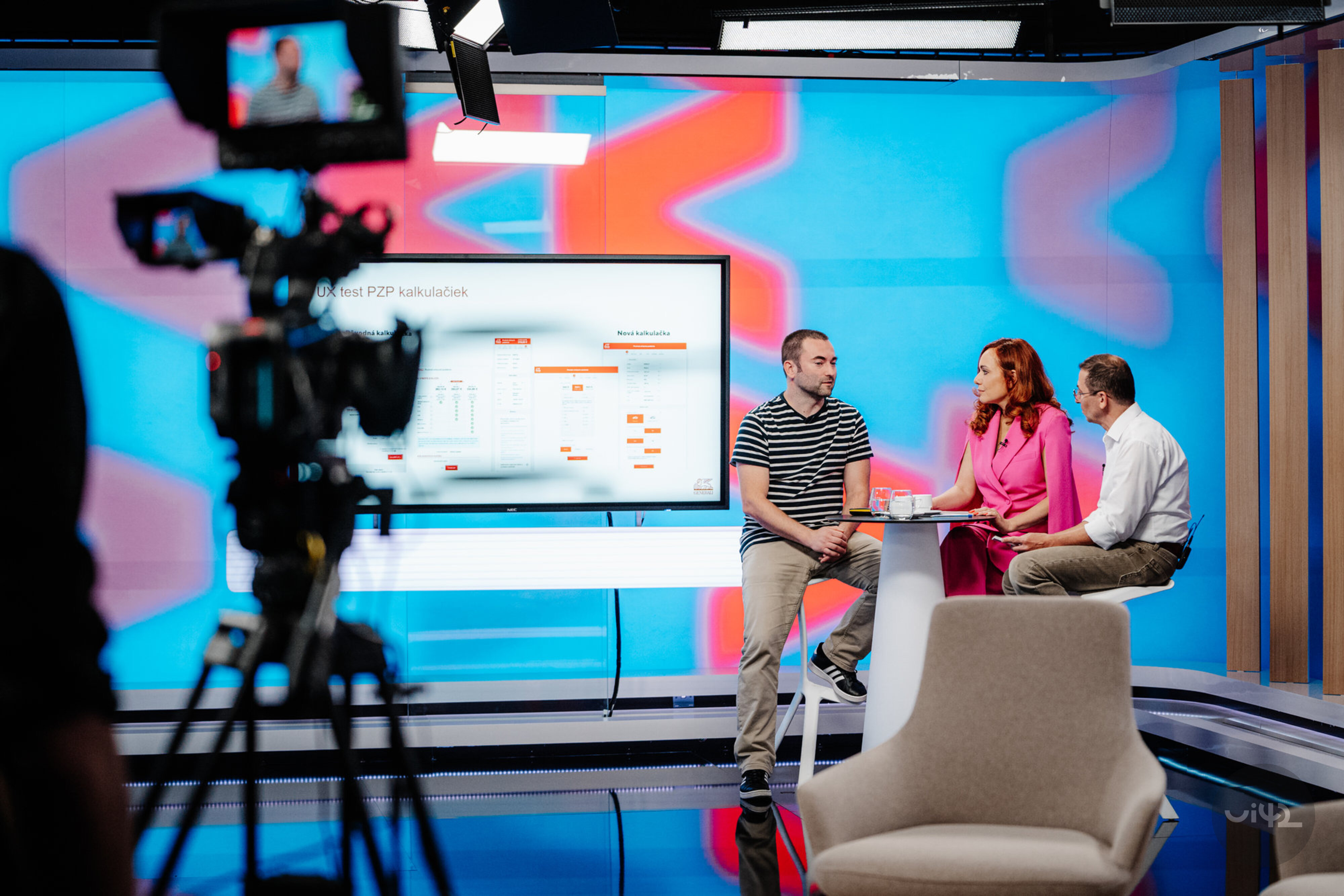 Diskusia v televíznom štúdiu so zameraním na UX testy PZP kalkulačiek. Traja ľudia sedia pri stole, pričom na obrazovke za nimi sú zobrazované grafy a údaje. Pred nimi je televízna kamera, ktorá ich sníma.