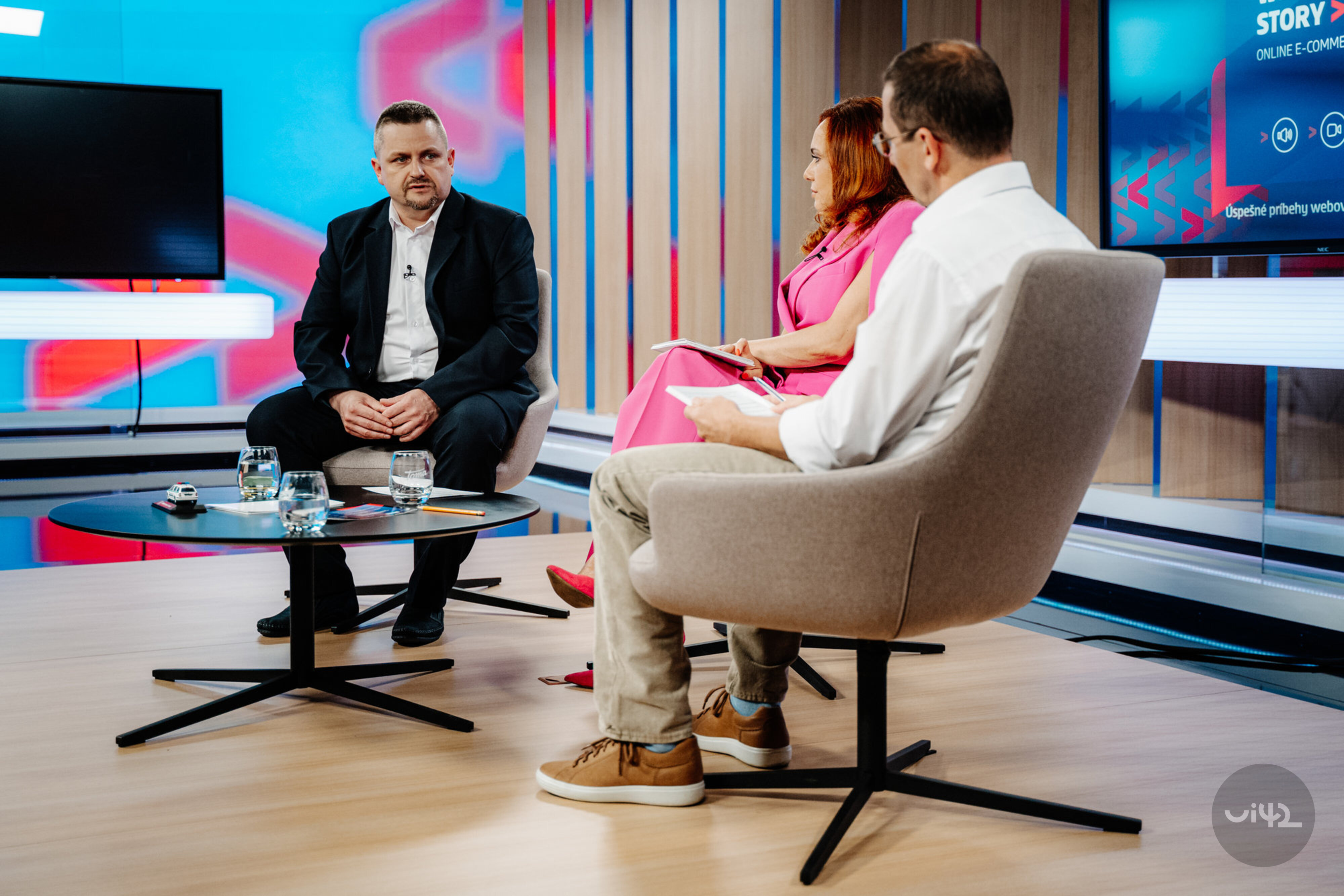 Diskusia troch ľudí v televíznom štúdiu, muž v obleku, žena v ružových šatách a muž v bielej košeli so svetlými nohavicami, sedia okolo stola s pohármi vody.