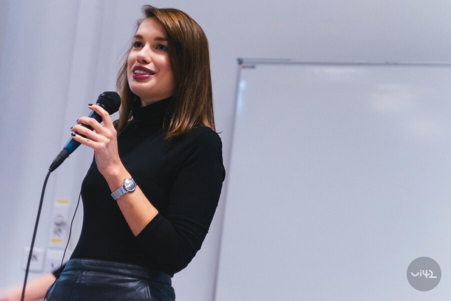 Žena prednáša na konferencii s mikrofónom v ruke, stojí pred bielou tabuľou. Má na sebe čierny rolák a hodinky na zápästí.