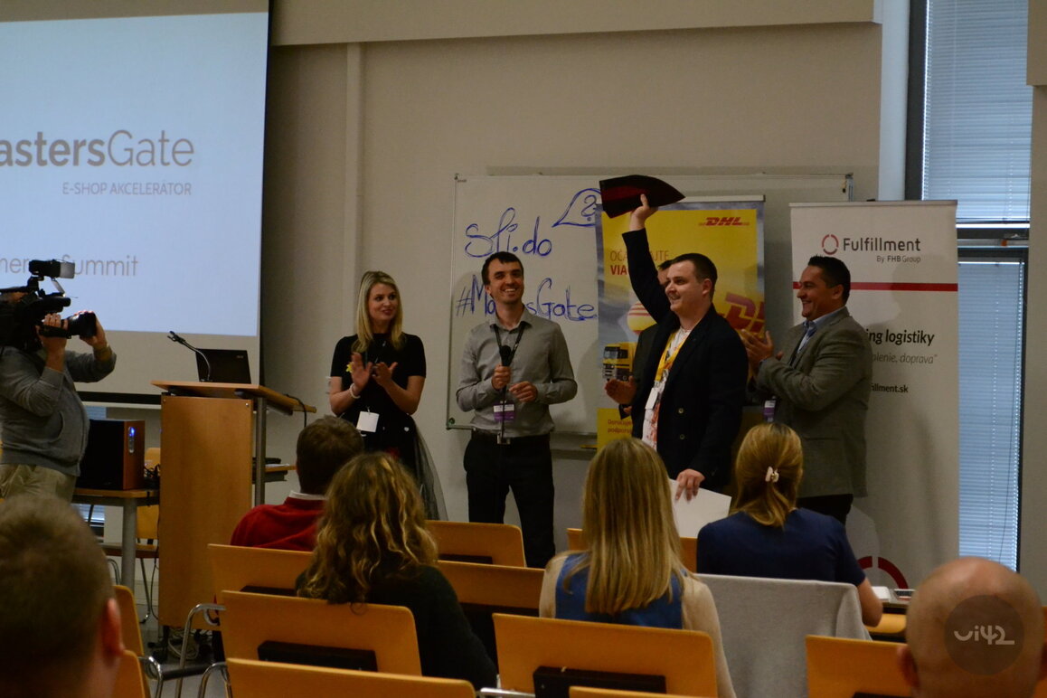 Ľudia na MastersGate E-commerce Summit počas prezentácie, muži a žena na pódiu, jeden muž dvíha ruku s predmetom, publikum sedí v miestnosti.