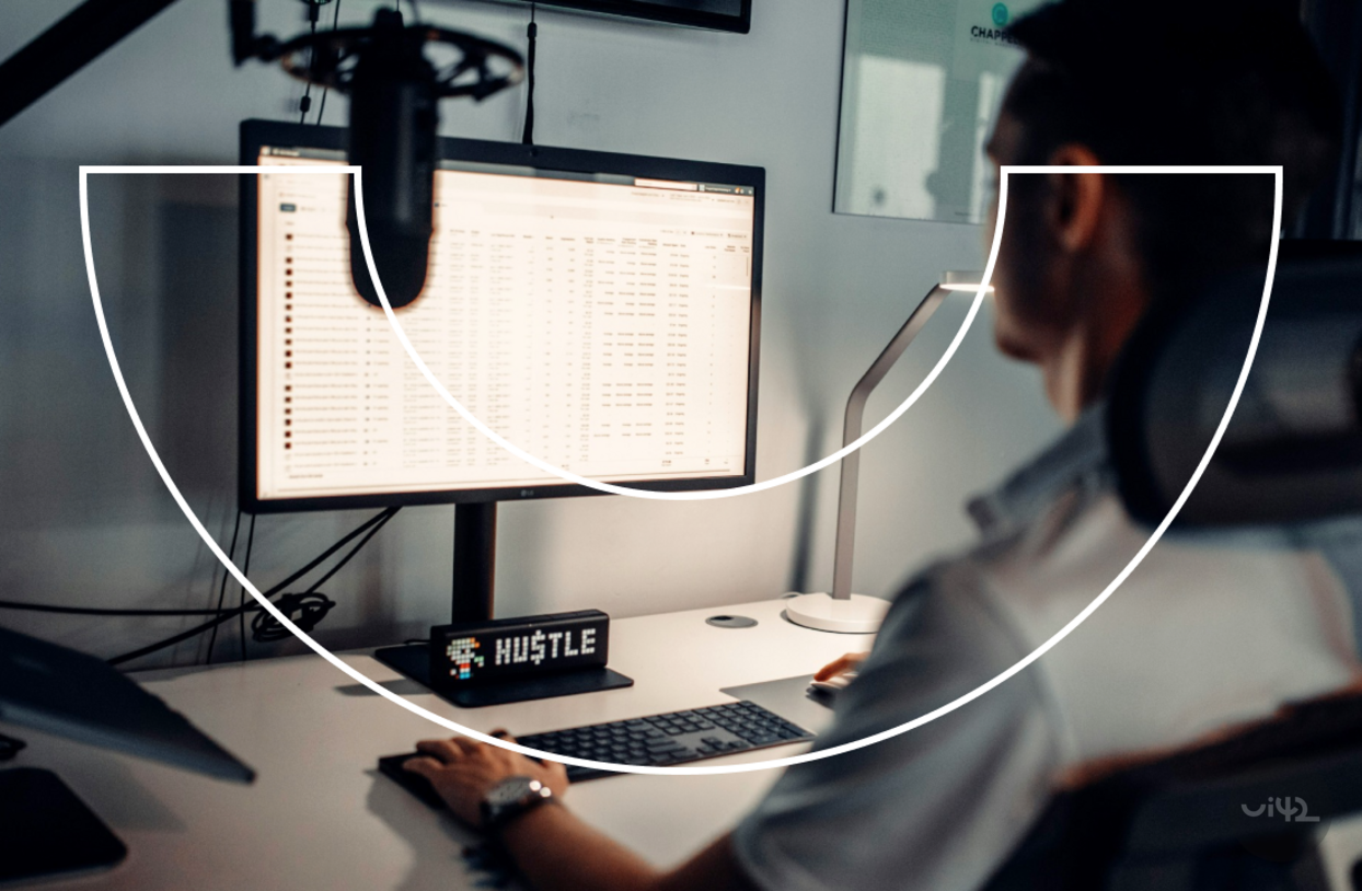 Osoba pracujúca pri počítači so zobrazenými dátami na monitore, na stole je nápis 'Hustle'.